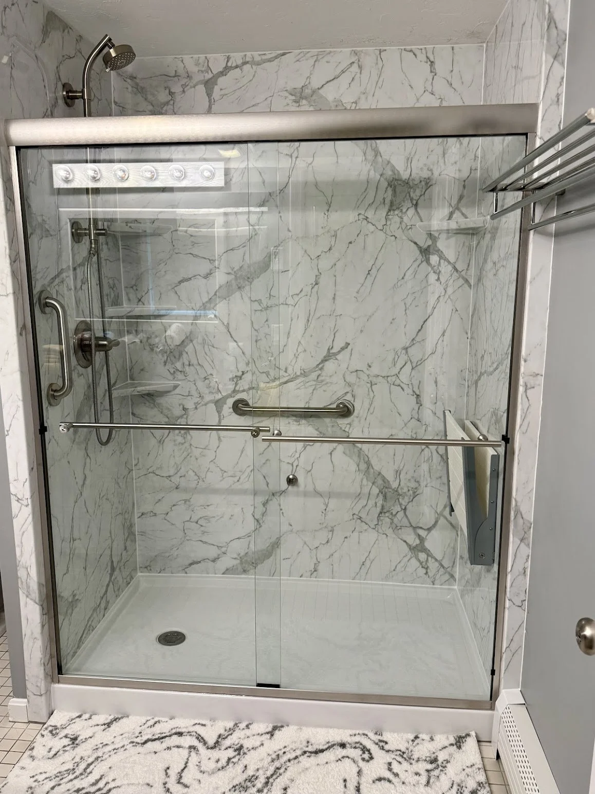 A completed walk-in shower installation in a South Shore Boston home, featuring elegant marble-look wall panels, a handheld showerhead, and brushed nickel fixtures. The space includes a grab bar, built-in shelving, and a sliding glass door system—delivering both accessibility and modern style. The transformation showcases Bay State Bath’s craftsmanship and attention to detail from design to final install.