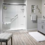 Modern bathroom with a walk-in shower featuring white subway tile walls, chrome grab bars, a hexagonal corner seat, and a clear glass sliding door. Room includes a gray vanity, light wood flooring, and decorative wall shelving.