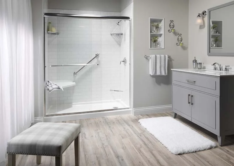 Modern bathroom with a walk-in shower featuring white subway tile walls, chrome grab bars, a hexagonal corner seat, and a clear glass sliding door. Room includes a gray vanity, light wood flooring, and decorative wall shelving.