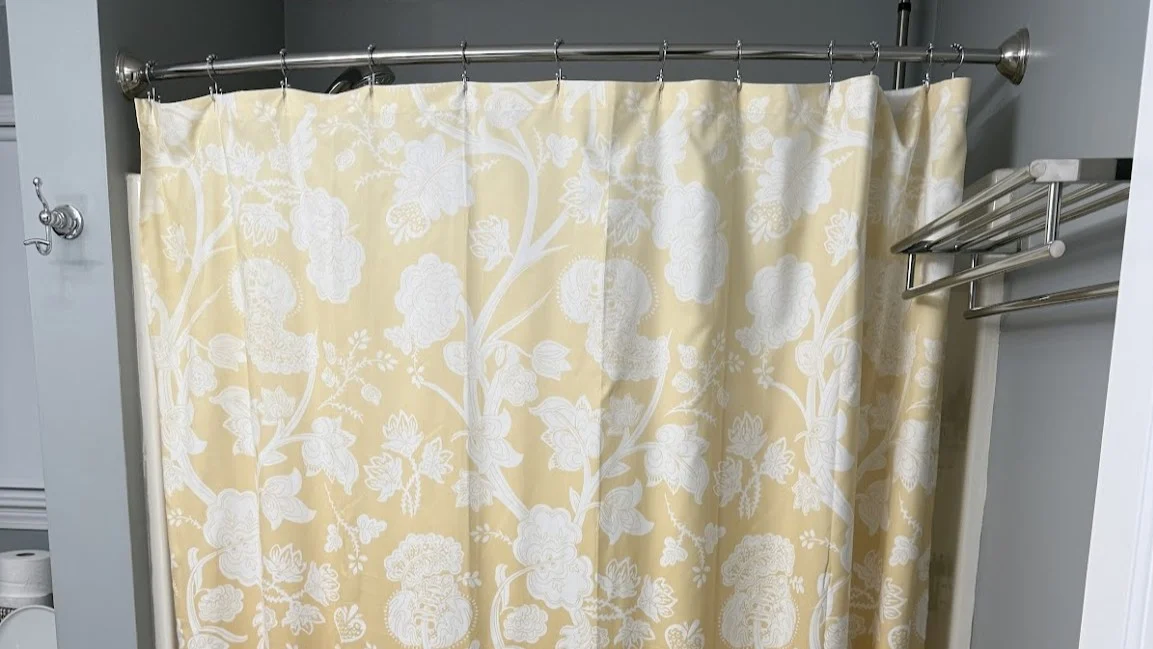 A “before” image of a traditional tub-and-shower setup in a South Shore Boston home. The space features a curved shower rod with a floral-patterned yellow and white curtain, light gray walls, and a metal towel rack. Though neat and functional, the setup lacks modern accessibility and visual appeal—setting the stage for a dramatic walk-in shower transformation.