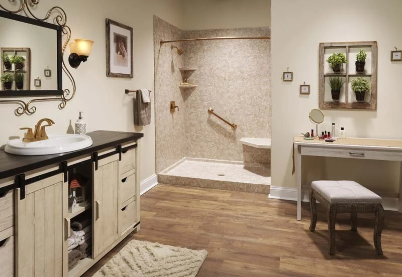 Warm-toned bathroom with a walk-in shower featuring a stone-look wall surround, bronze fixtures, grab bars, and a corner seat. The room includes rustic wood cabinetry, a makeup vanity, wood-style flooring, and decorative greenery.