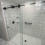 A newly remodeled walk-in shower in a South Shore Boston home featuring large subway-style wall panels, matte black hardware, recessed shelving, and a textured white accent wall. The frameless glass doors and marble-look tile flooring highlight Bay State Bath’s clean, high-end installation style.
