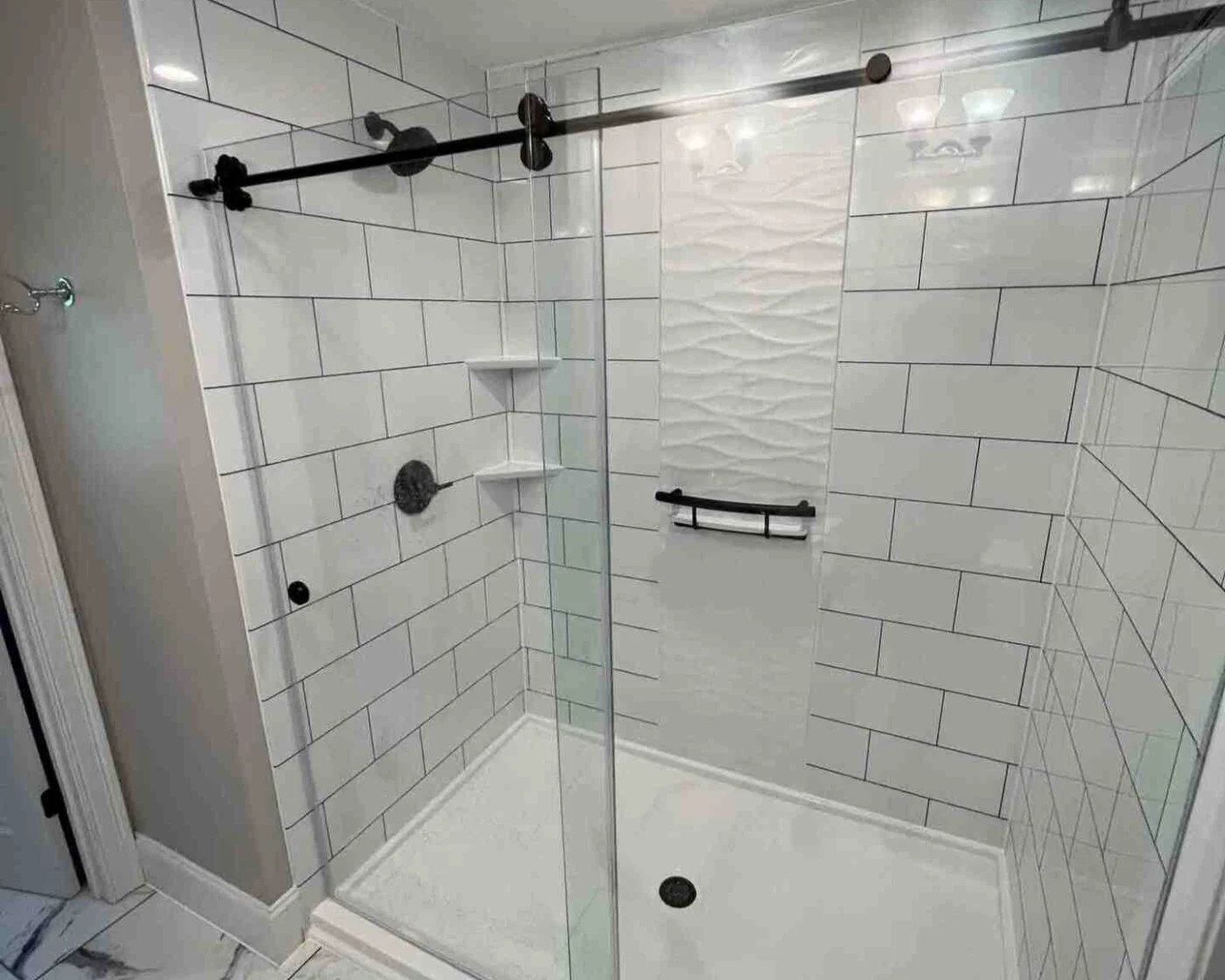 A newly remodeled walk-in shower in a South Shore Boston home featuring large subway-style wall panels, matte black hardware, recessed shelving, and a textured white accent wall. The frameless glass doors and marble-look tile flooring highlight Bay State Bath’s clean, high-end installation style.