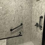 Walk-in shower installed by Bay State Bath with striking marble-look wall surround, accented by matte black fixtures, three grab bars, two built-in corner shelves, and a clear glass door with black trim.