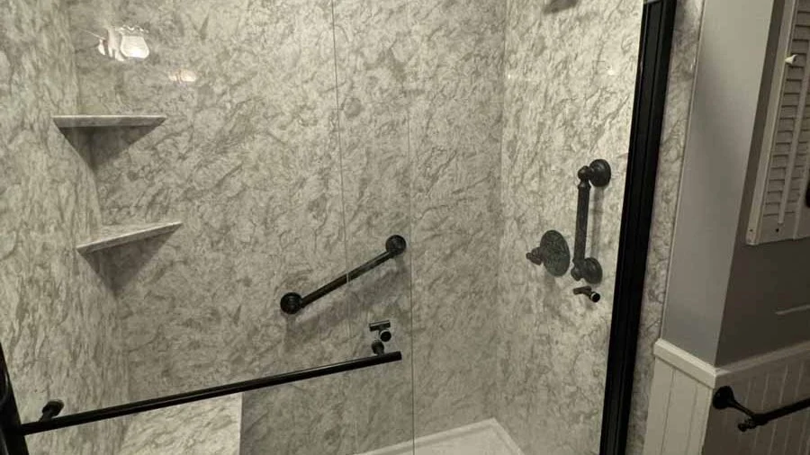 Walk-in shower installed by Bay State Bath with striking marble-look wall surround, accented by matte black fixtures, three grab bars, two built-in corner shelves, and a clear glass door with black trim.