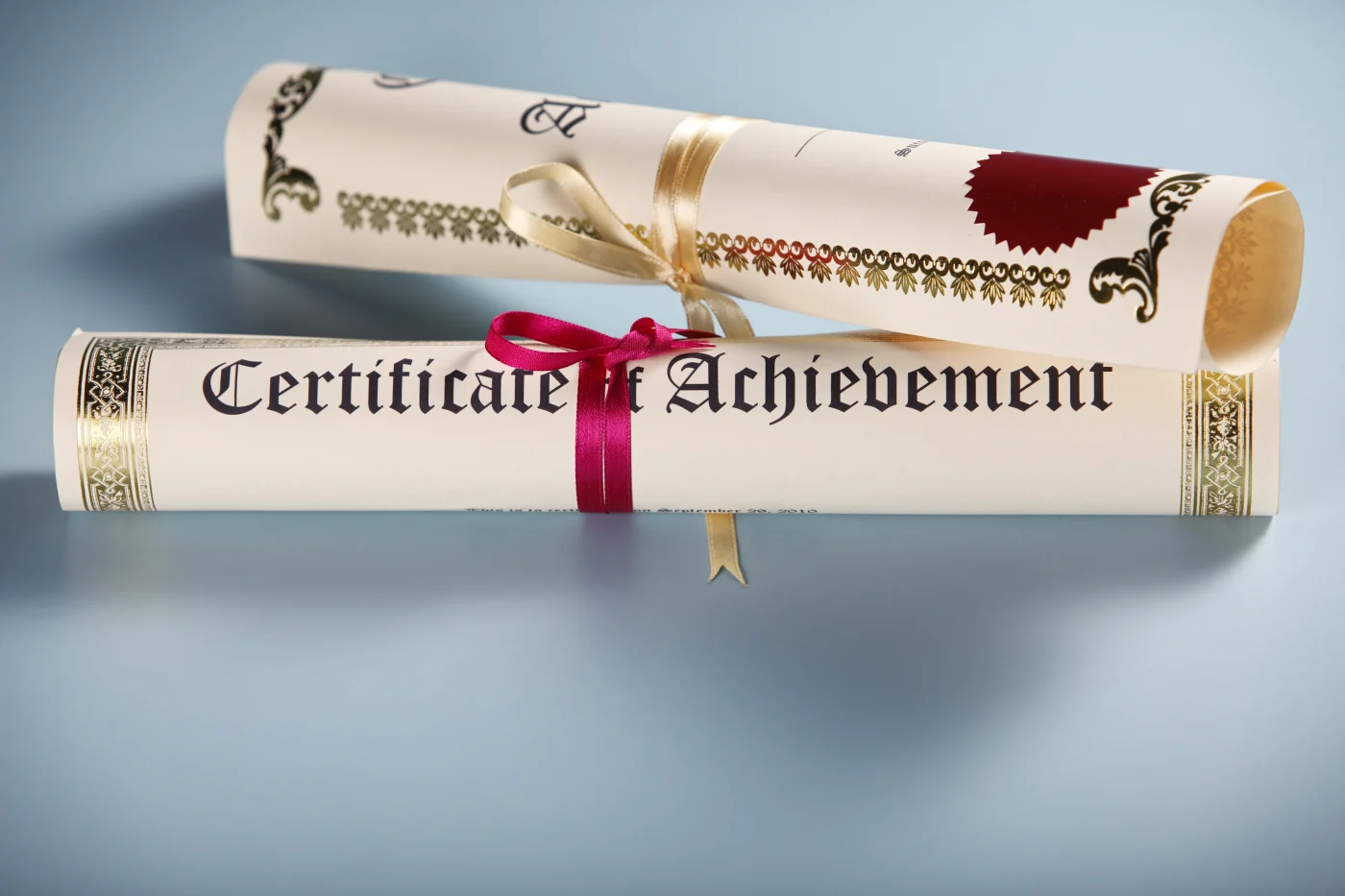 Two rolled up certificates with red and gold ribbon. One reads certificate of achievement