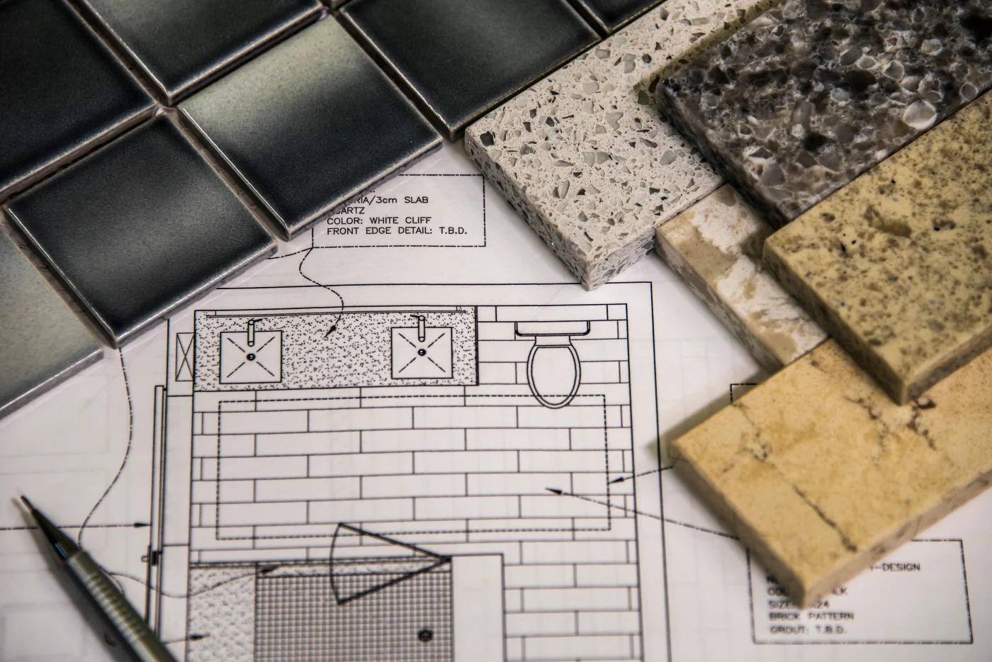 A variety of bathroom countertop and tile samples are arranged on top of a detailed floor plan sketch. The samples include different colors and textures, from dark ceramic tiles to light and speckled quartz and stone finishes. A pen rests nearby, suggesting active planning and design. The image conveys a personalized, hands-on approach to bathroom remodeling and material selection.