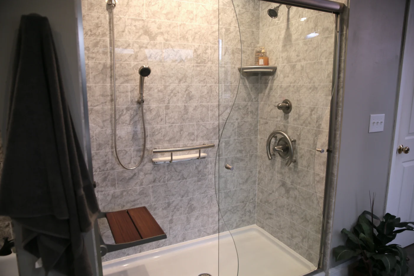 A walk-in shower with a glass door, white base, gray wall, silver features, and a wood seat.