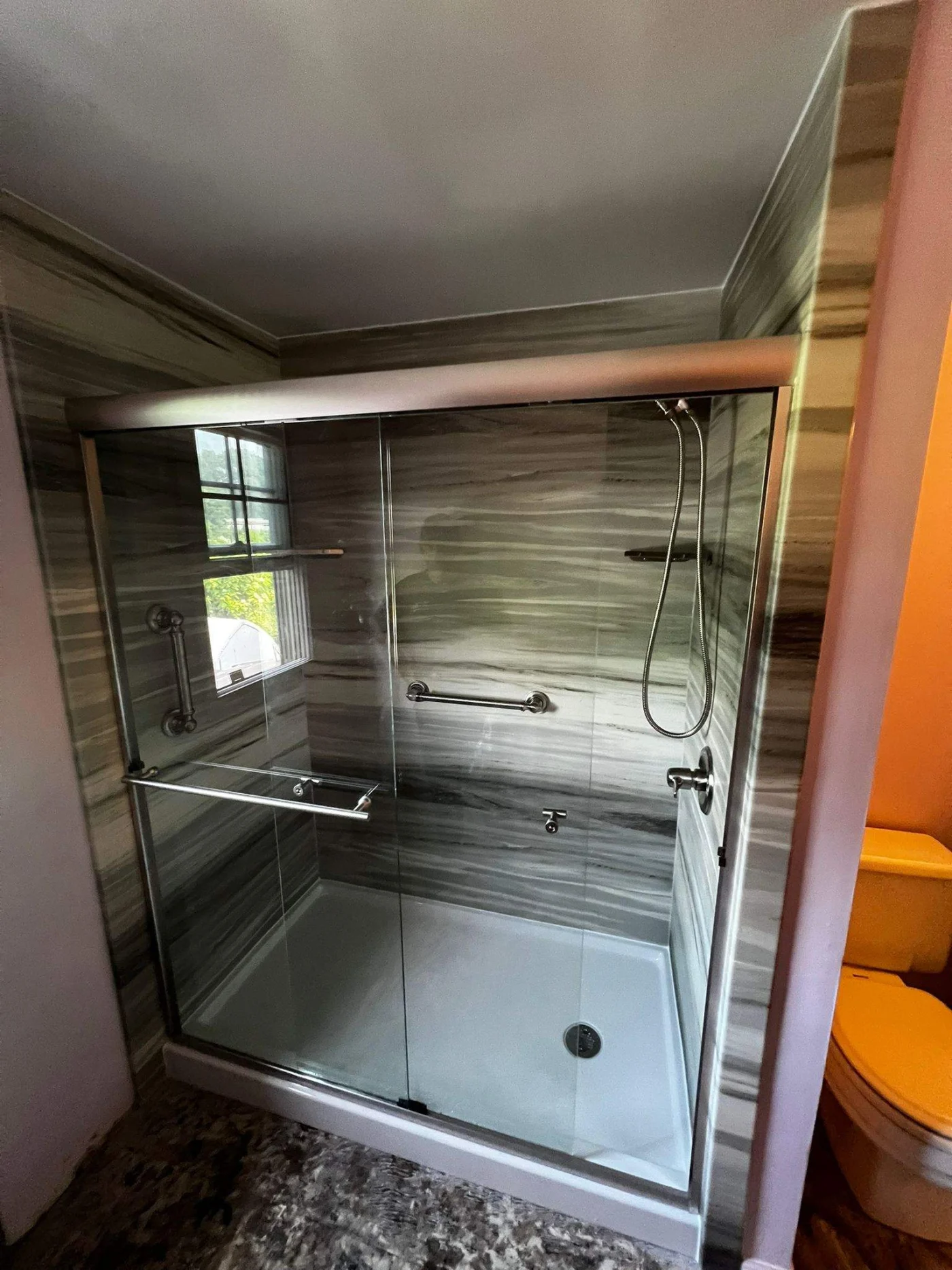A walk-in shower with a glass door, white base, and gray wall with silver features.