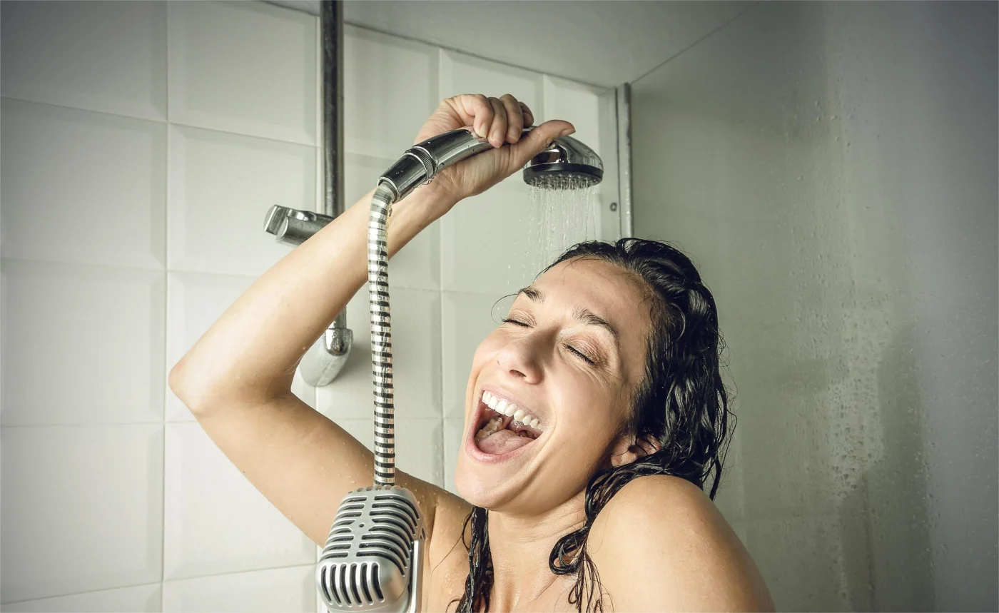 A woman singing in the shower with a shower head over her and a microphone