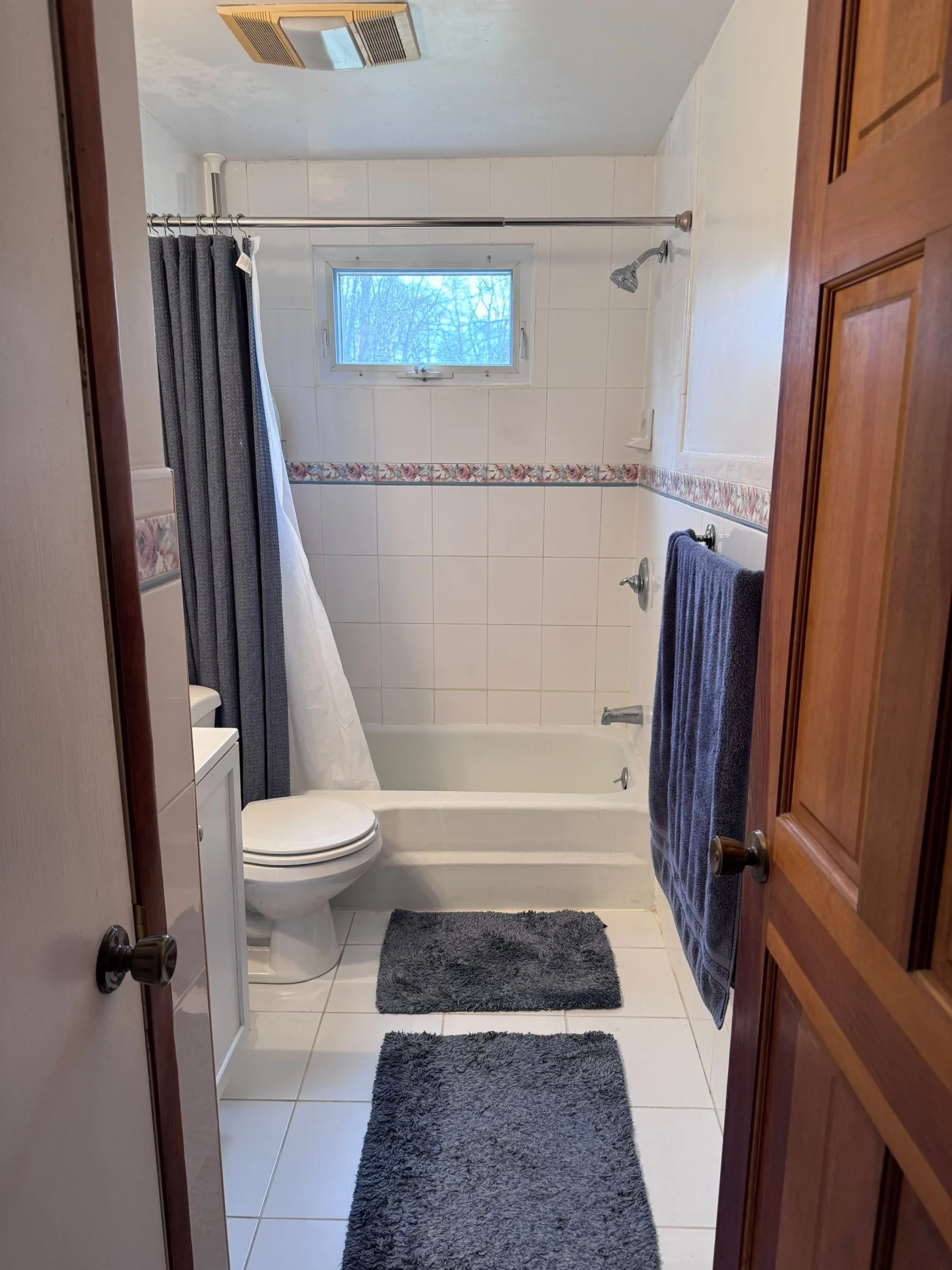 A white bathroom with white flooring and a white tub-shower combo with silver fixtures and a window.