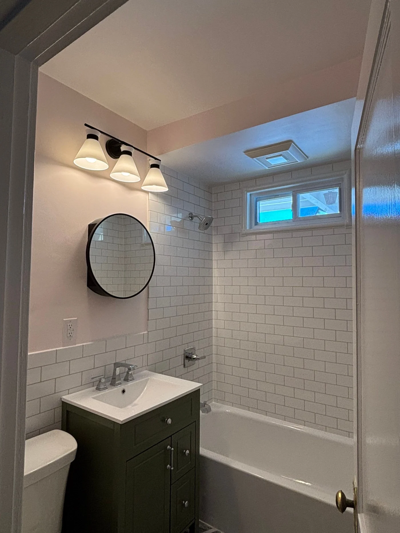 A white bathroom with a brown vanity, a white sink, a circular mirror, overhead lighting, and a white bathtub.