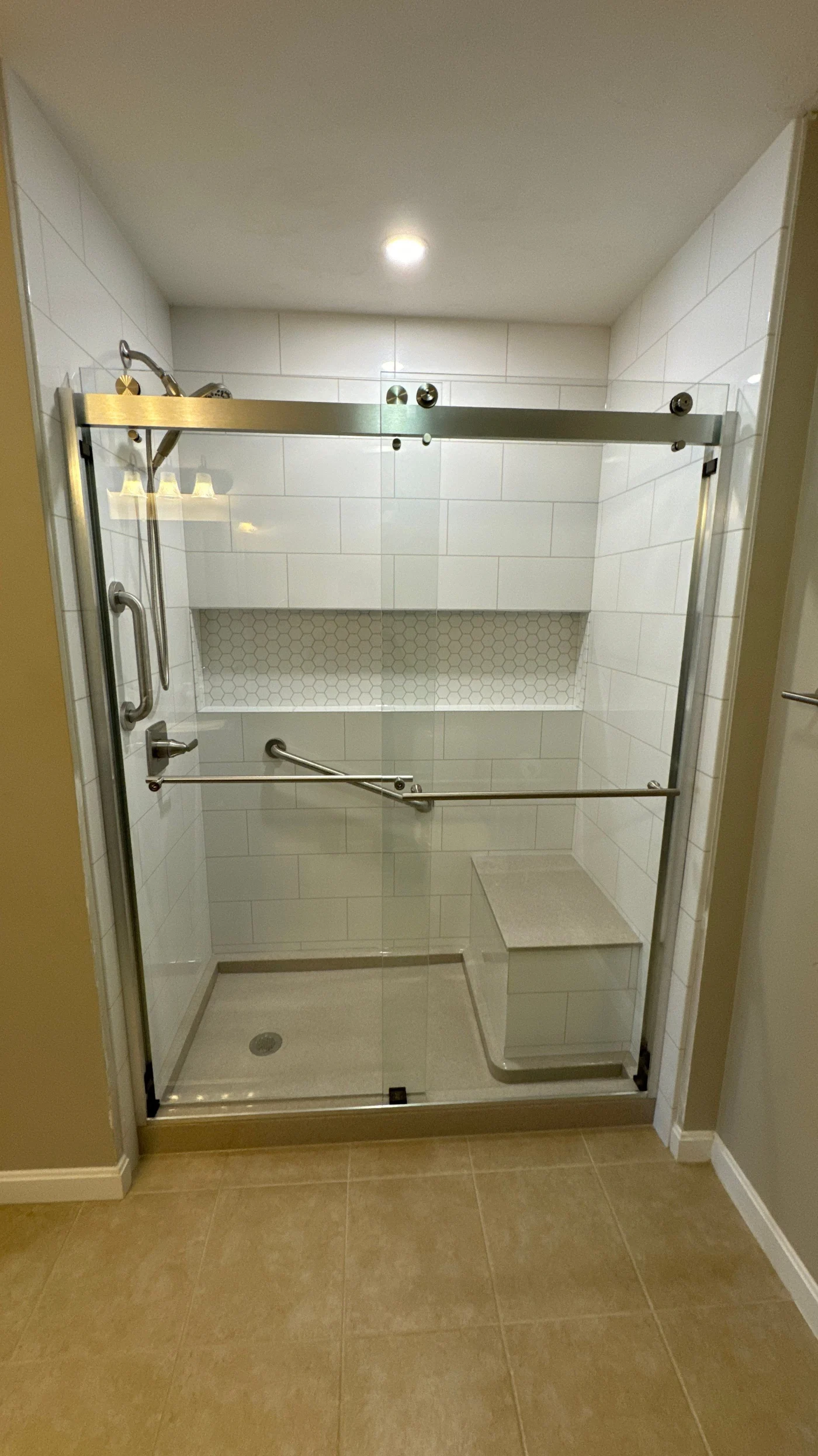 A white walk-in shower with a gray base, a white bench, silver fixtures, and a sliding glass door.