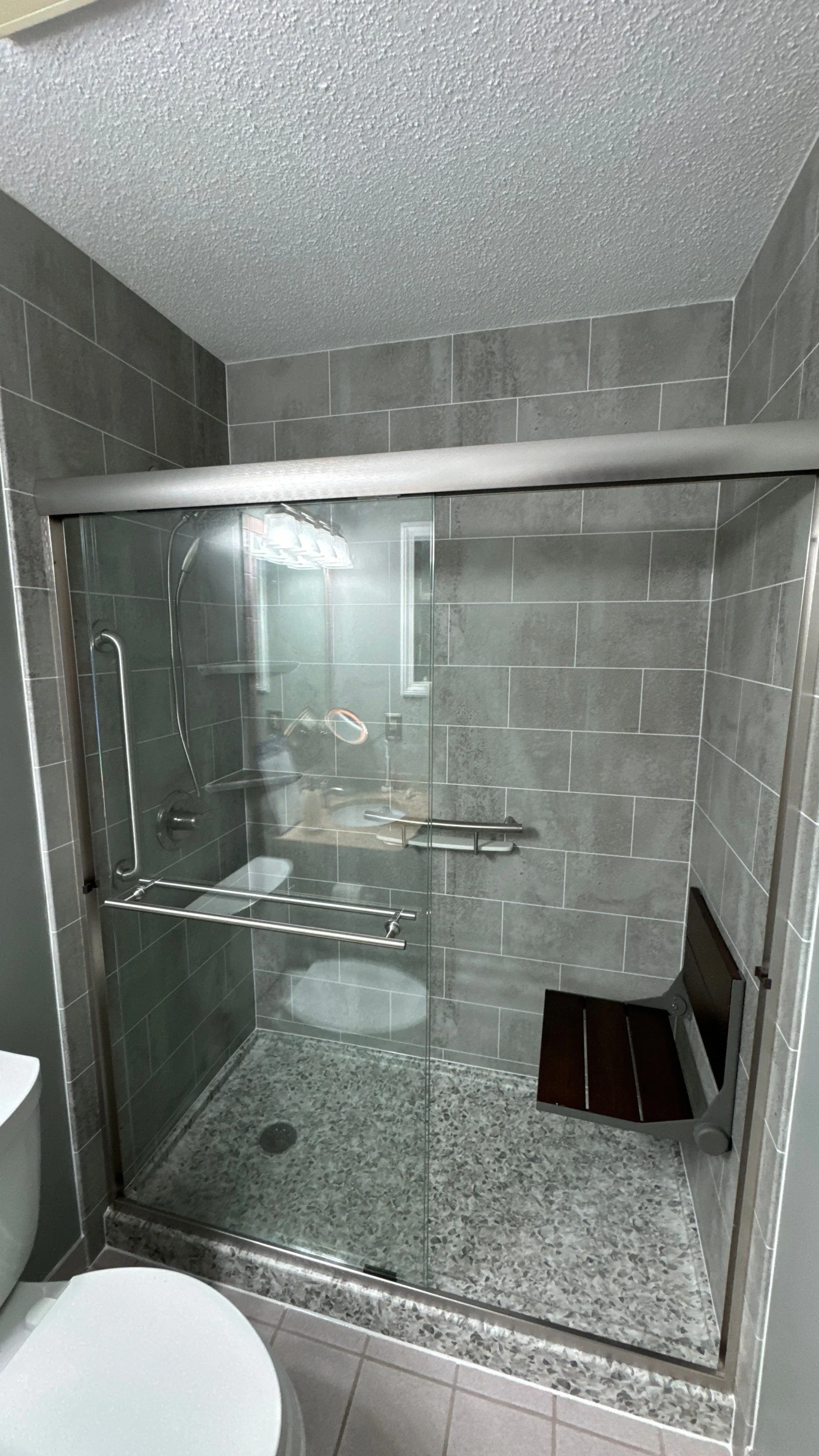 A gray walk-in shower with a gray base, a black seat, silver fixtures, and a sliding glass door.