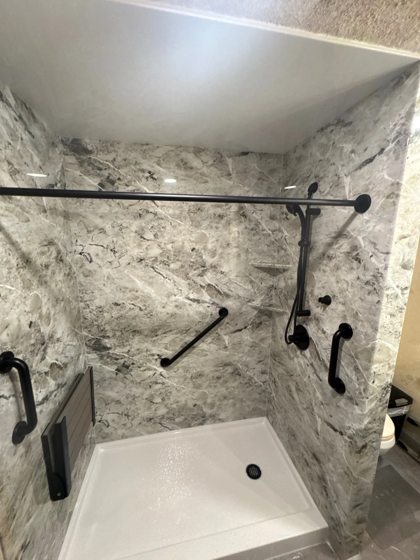 A walk-in shower with gray walls, a fold-up seat, black fixtures, and a white base.