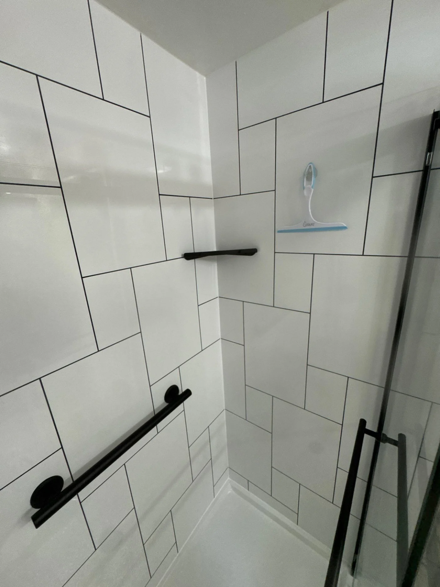 A close-up view of a white shower wall with a black hand rail and shelf in the corner.