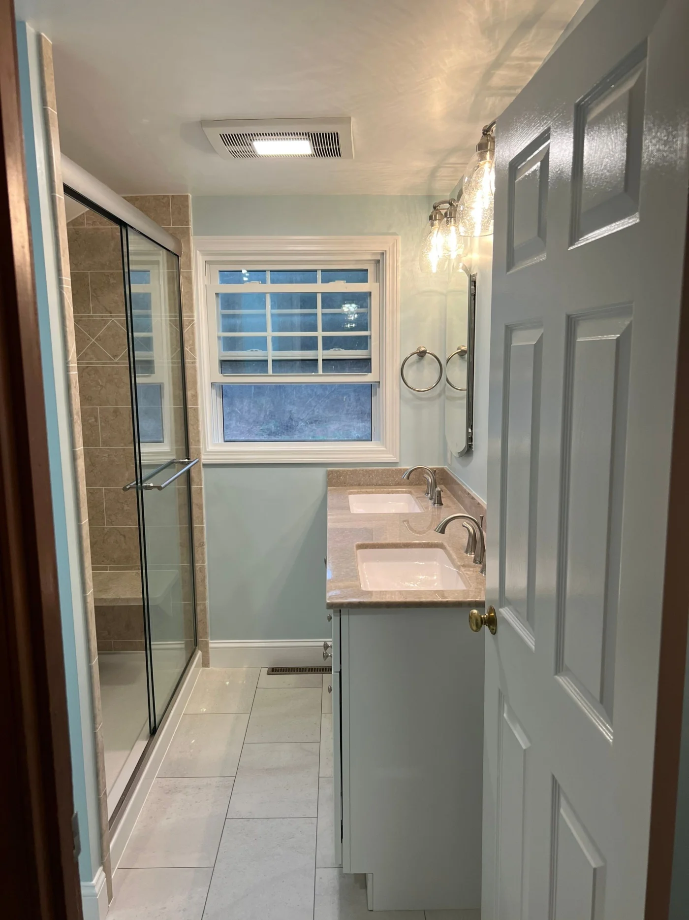 A blue bathroom with a blue vanity, two sinks, and a walk-in shower with a window at the end.