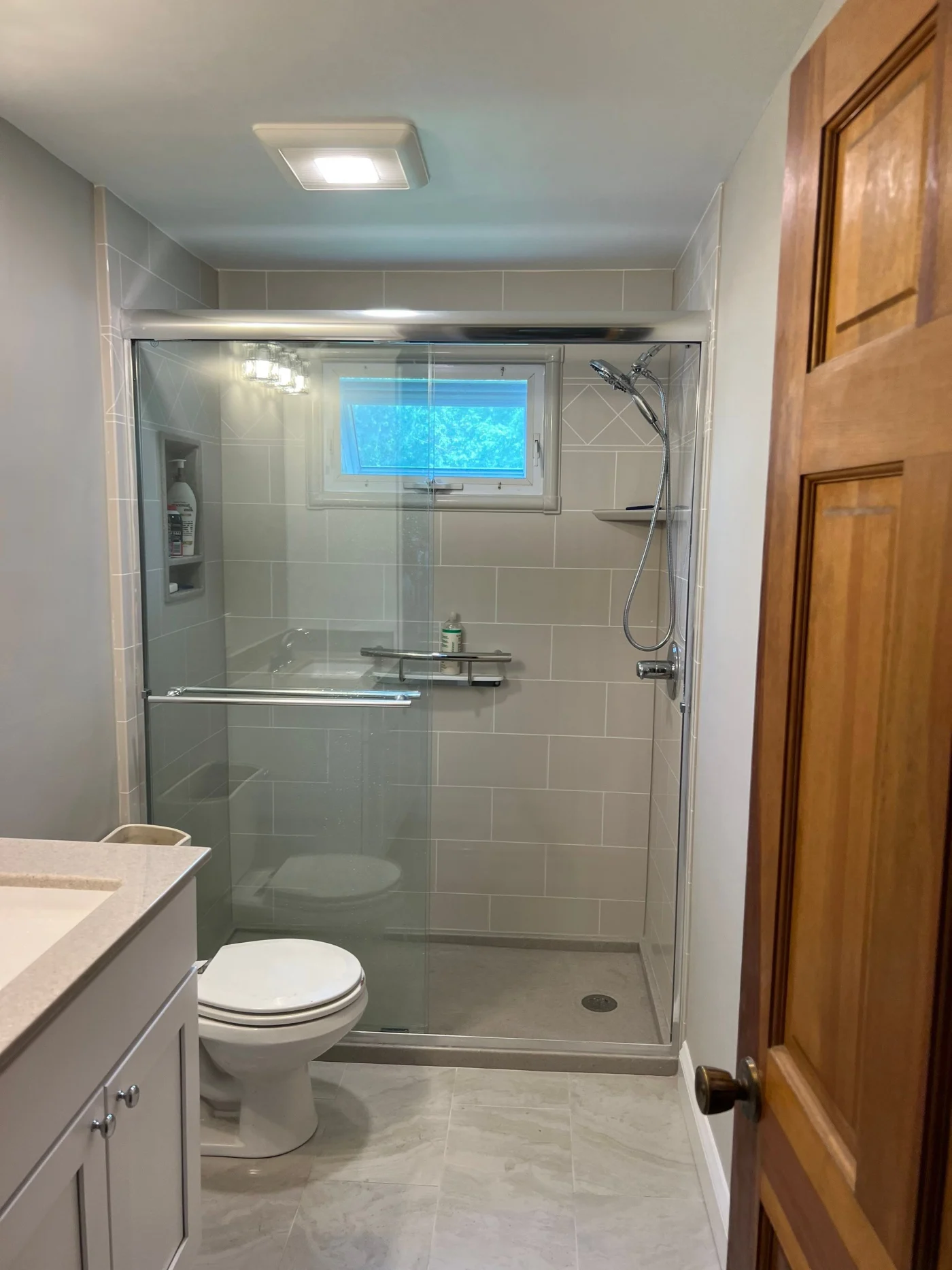 A white bathroom with a gray walk-in shower featuring silver fixtures, a sliding glass door, and a window.