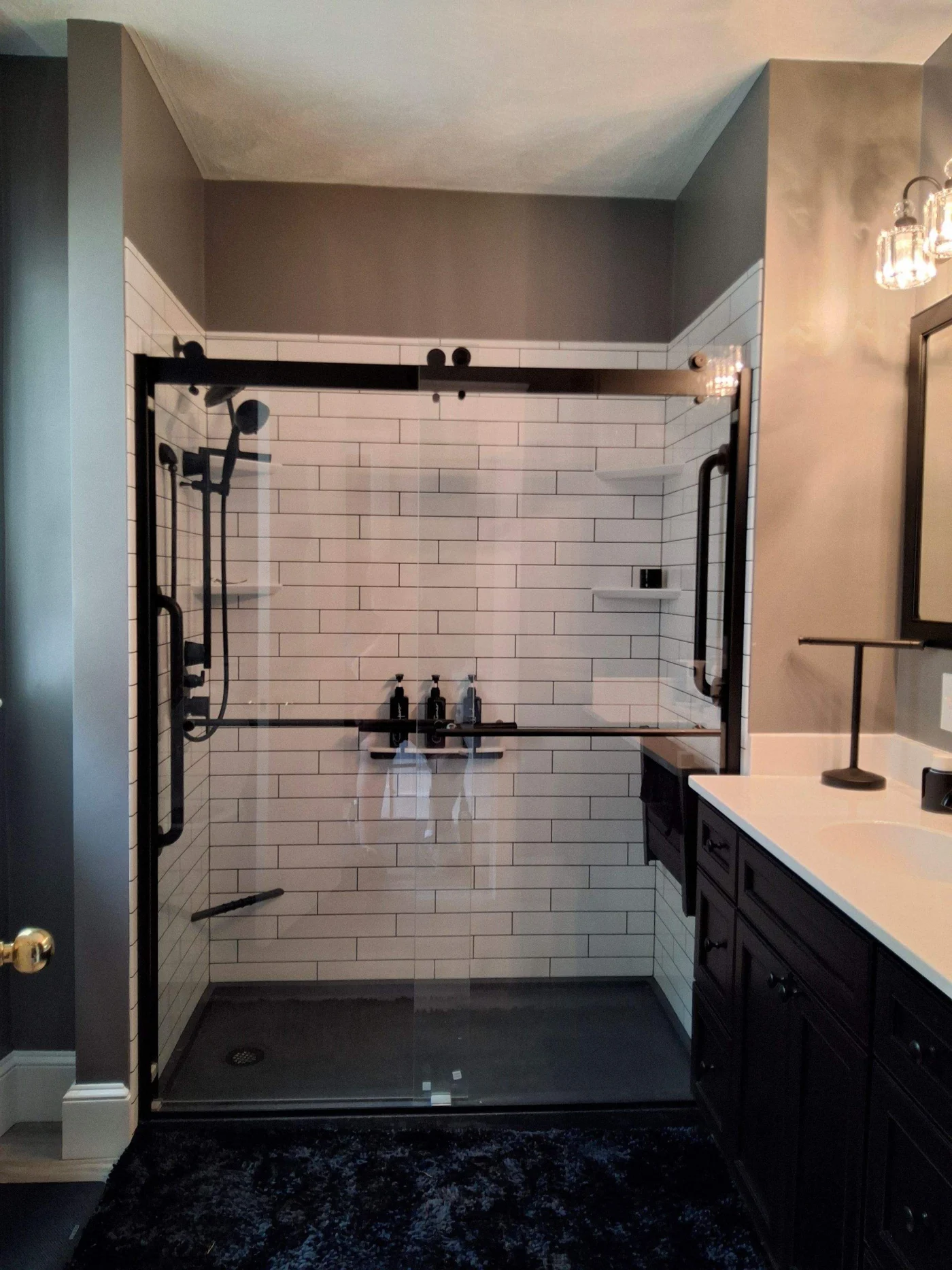 A gray bathroom with a black vanity, a white countertop, and a white walk-in shower with black fixtures and a glass door.