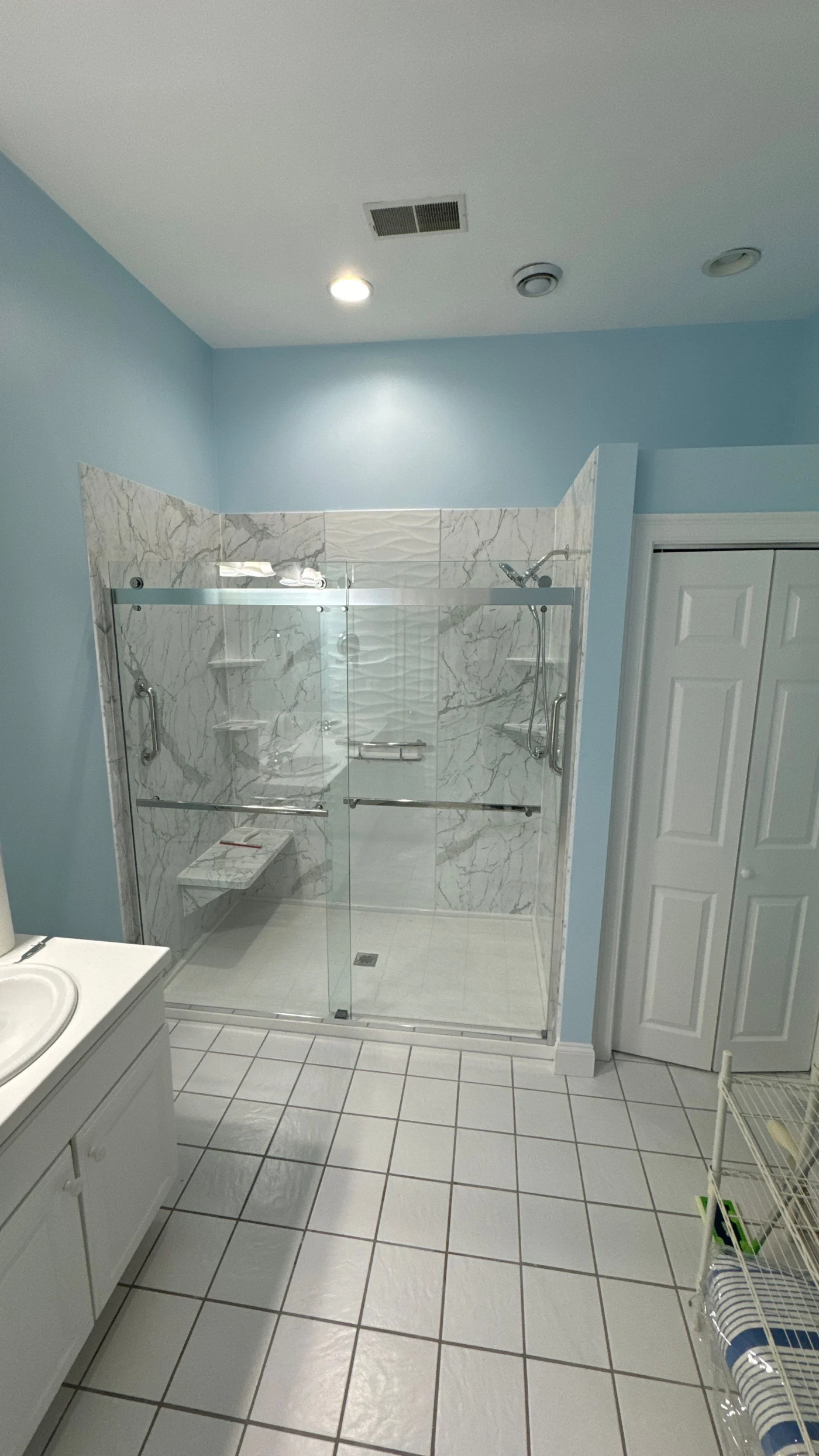 This bright full bathroom remodel features light blue walls, square floor tiles, and a spacious walk-in shower enclosed in glass with elegant marble-patterned wall panels and a built-in bench.