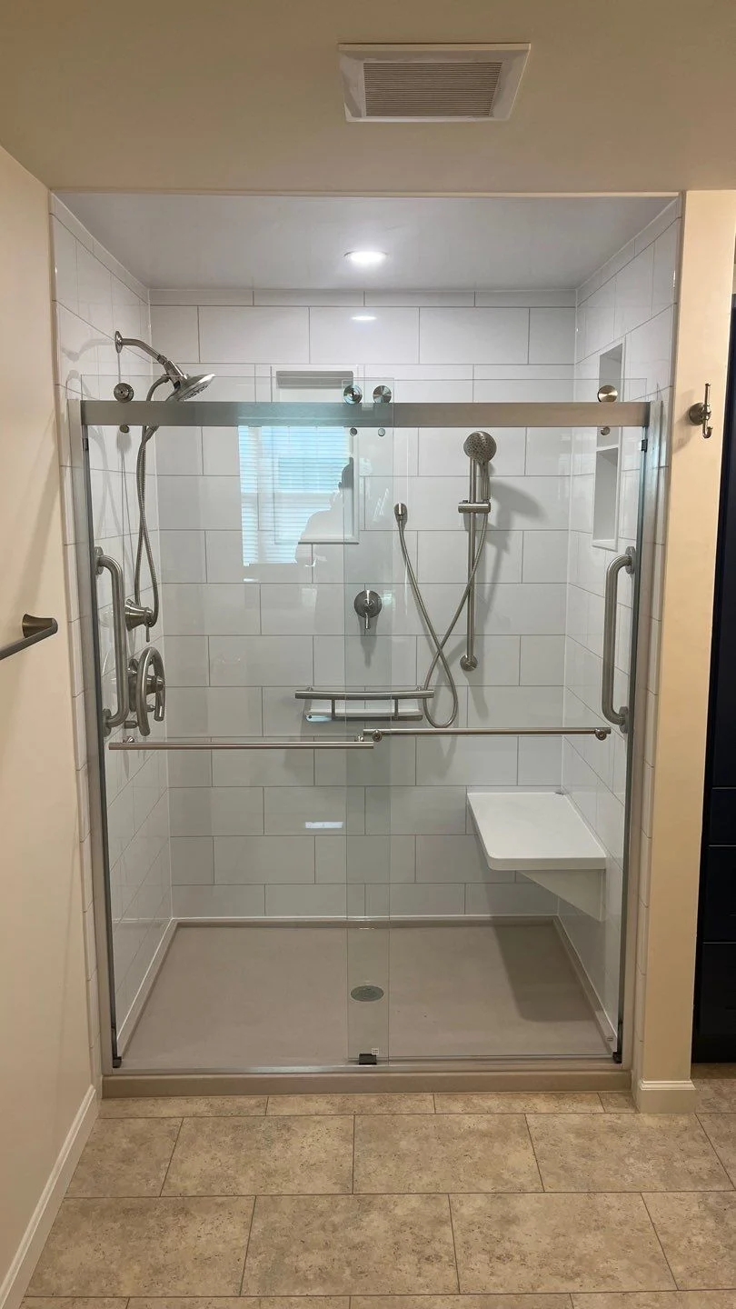 A walk-in shower with white walls, a gray base, silver fixtures, a white bench, and a sliding glass door.