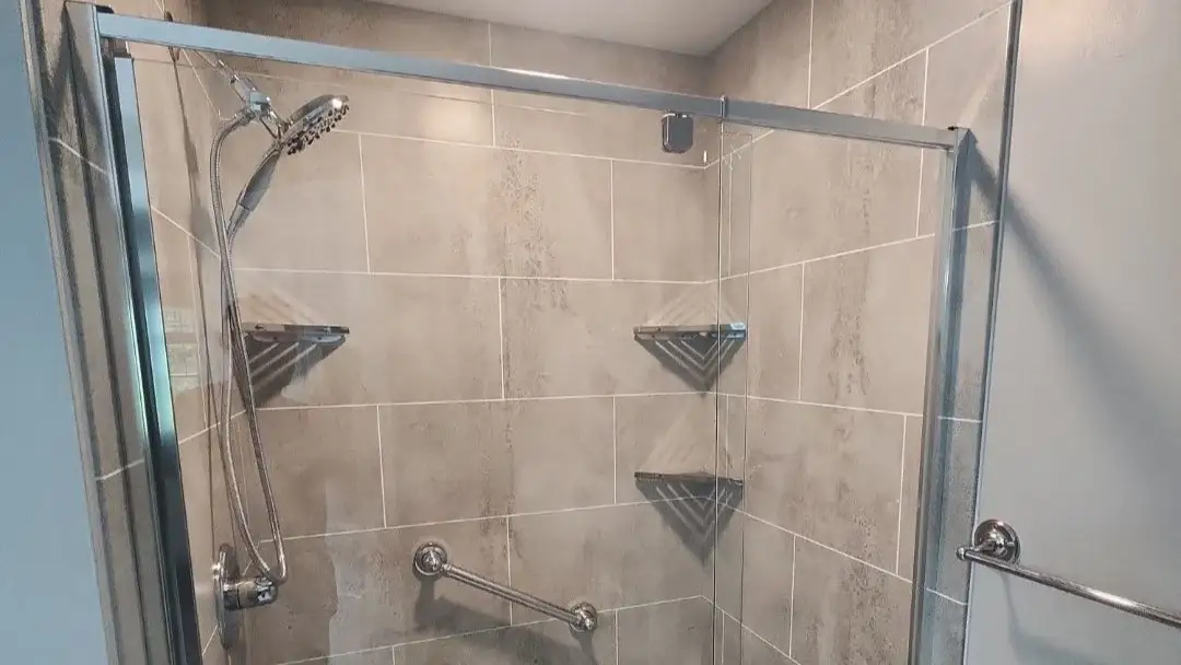 Walk-in shower with gray tile walls, glass doors, corner shelves, handheld showerhead, and grab bar.