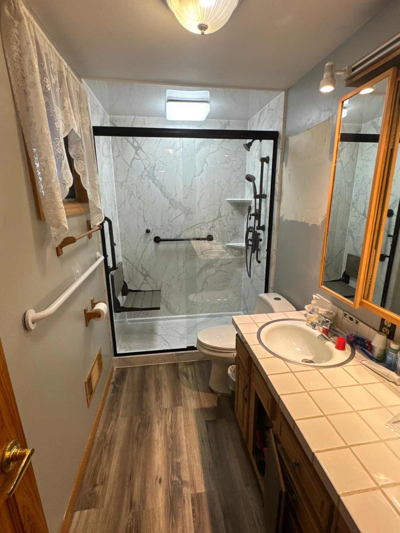 A long, narrow bathroom features wood-look flooring, a tiled vanity with a drop-in sink, and a black-framed glass walk-in shower with white marbled walls, a fold-down bench, and matte black fixtures.