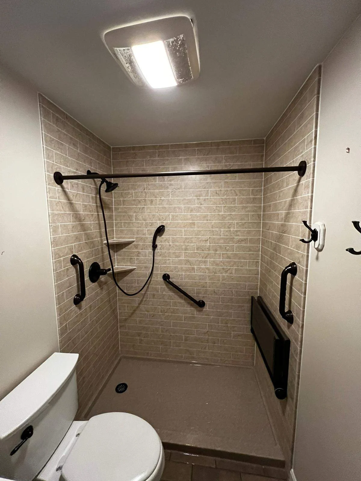 This accessible bathroom remodel features a barrier-free walk-in shower with neutral tan subway tile, matching corner shelves, matte black hardware, and several strategically placed safety grab bars.