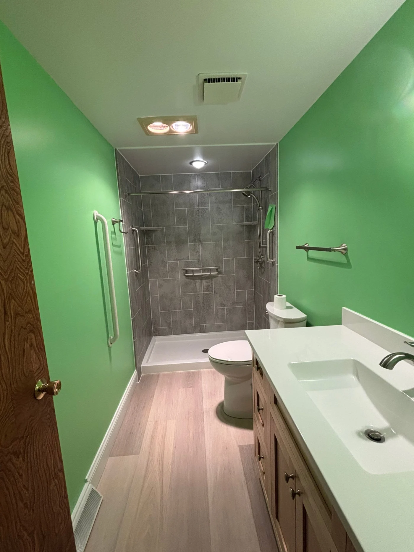 A long, narrow bathroom features vibrant lime green walls, a light wood vanity with a modern white sink, light wood-look flooring, and a walk-in shower lined with large gray horizontal tiles.