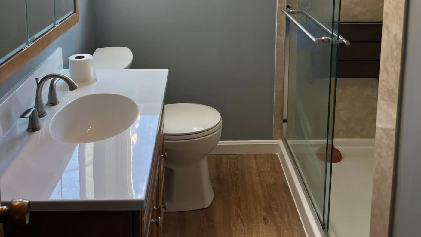 Remodeled bathroom with a glass-enclosed shower, white vanity, gray walls, and wood-look flooring.