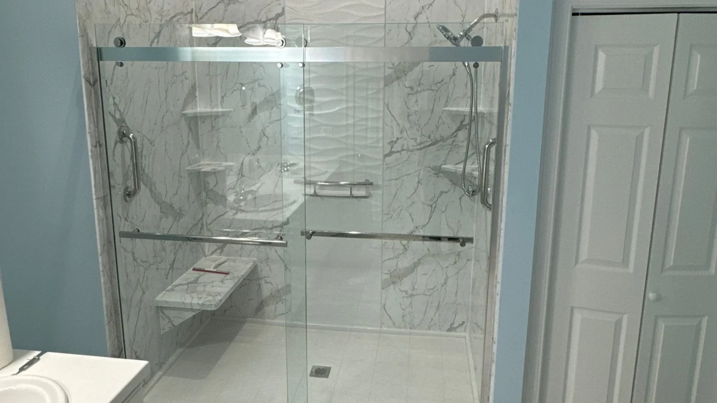 Spacious walk-in shower with marble-look walls, sliding glass doors, built-in bench, grab bars, and handheld showerhead.