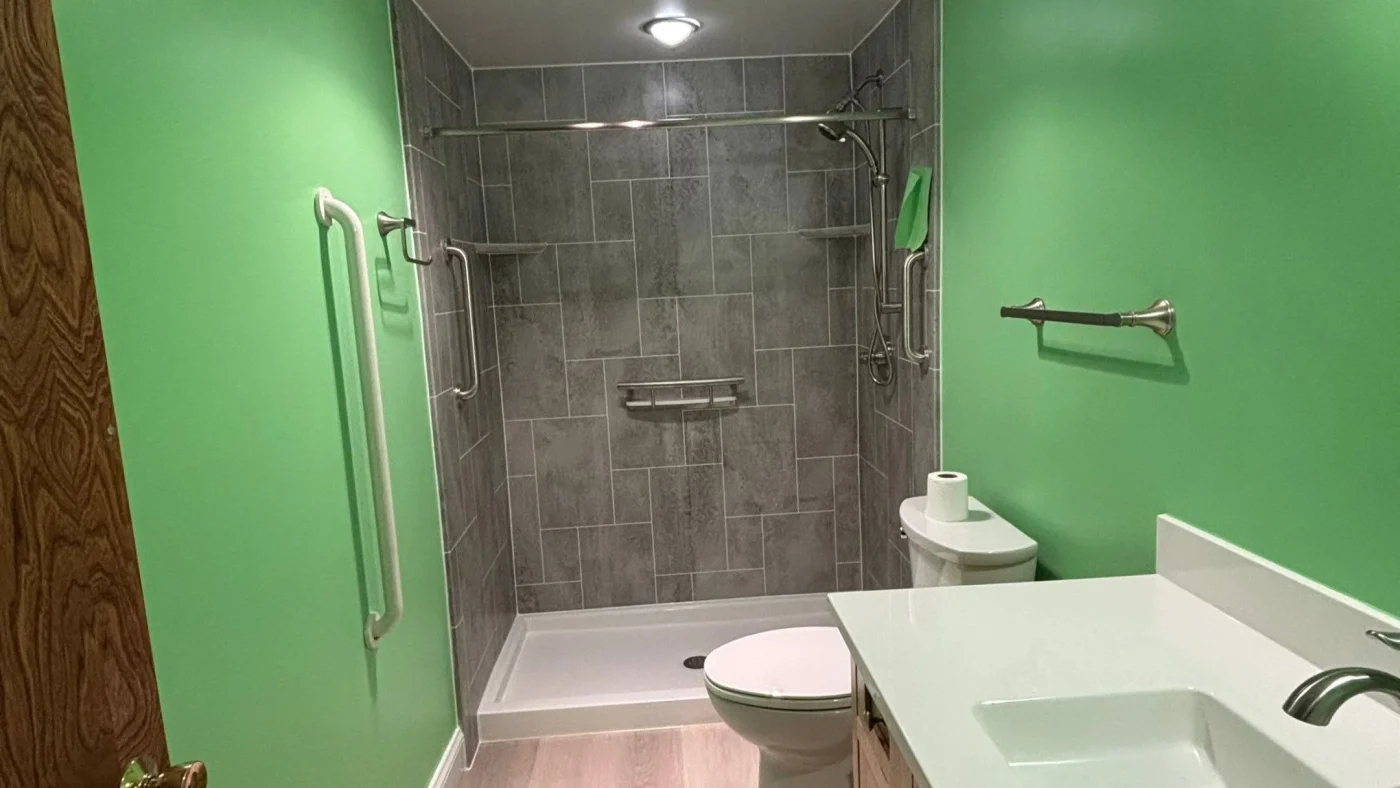 Remodeled bathroom with green walls, a gray tiled walk-in shower, grab bars, and a white vanity.