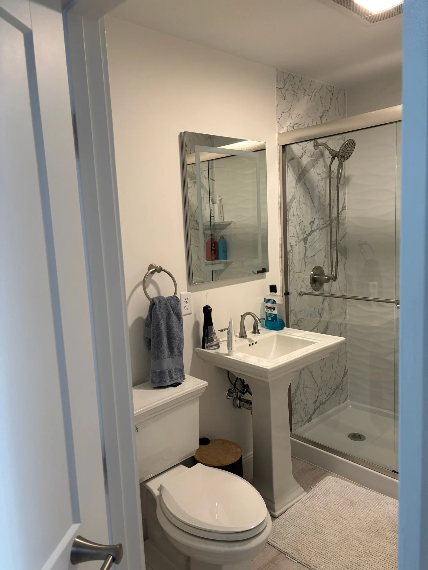 A white bathroom with a white toilet, a freestanding sink below a mirror, and a walk-in shower with a gray surround and a sliding glass door.