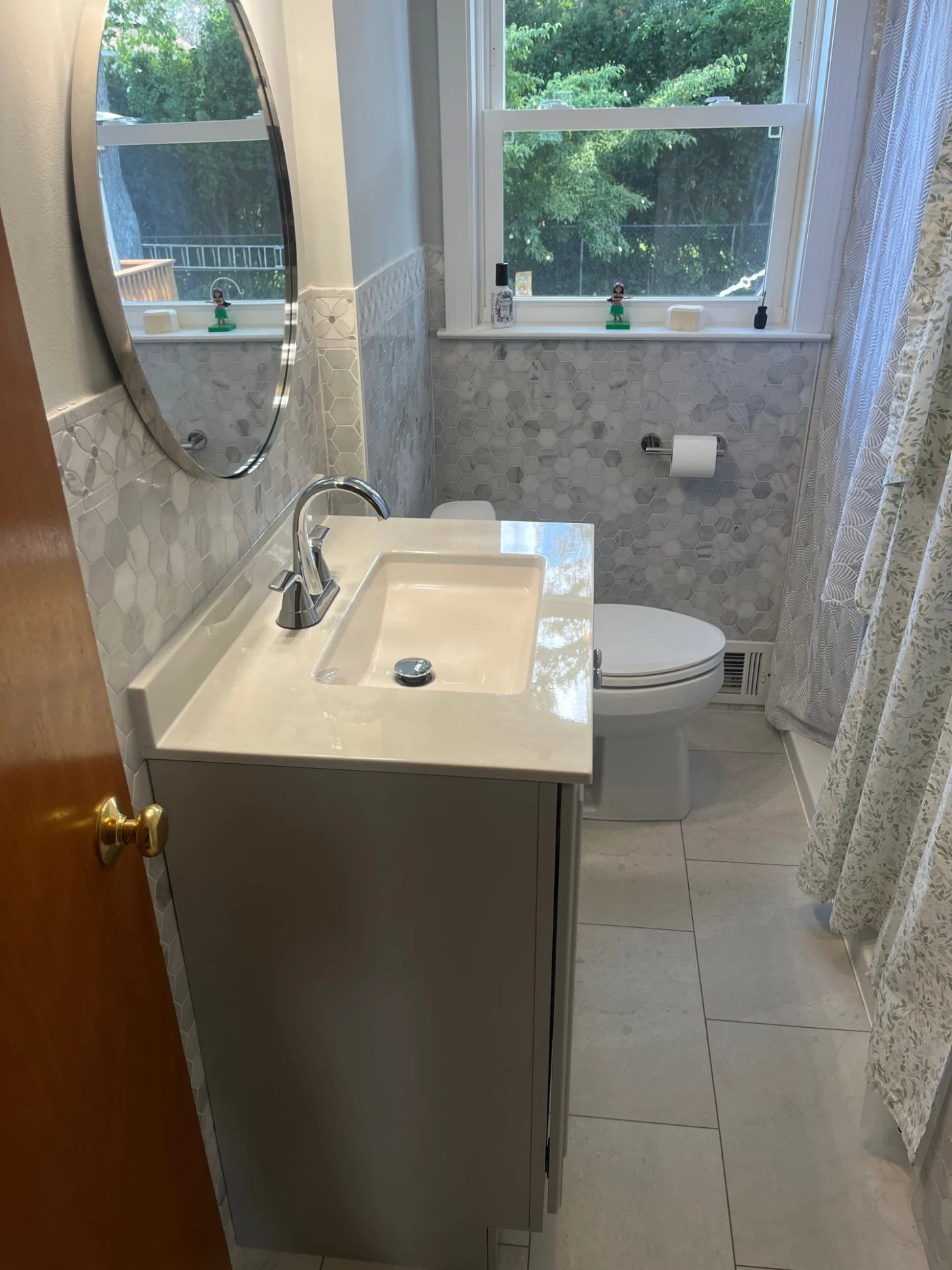 This stylish bathroom remodel highlights a grey single-sink vanity with a large oval mirror, beautifully paired with grey and white hexagonal mosaic wall tiles that extend beneath a bright window.