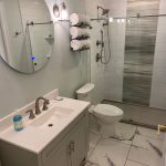 A bathroom with tile floors, a black vanity with a white sink and circular mirror, white walls, a white toilet, and a walk-in shower with a brown base, white and gray walls, and a glass door