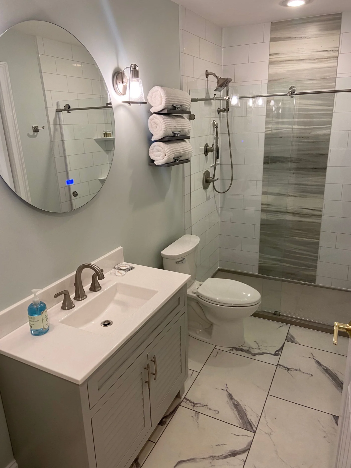 A bathroom with tile floors, a black vanity with a white sink and circular mirror, white walls, a white toilet, and a walk-in shower with a brown base, white and gray walls, and a glass door