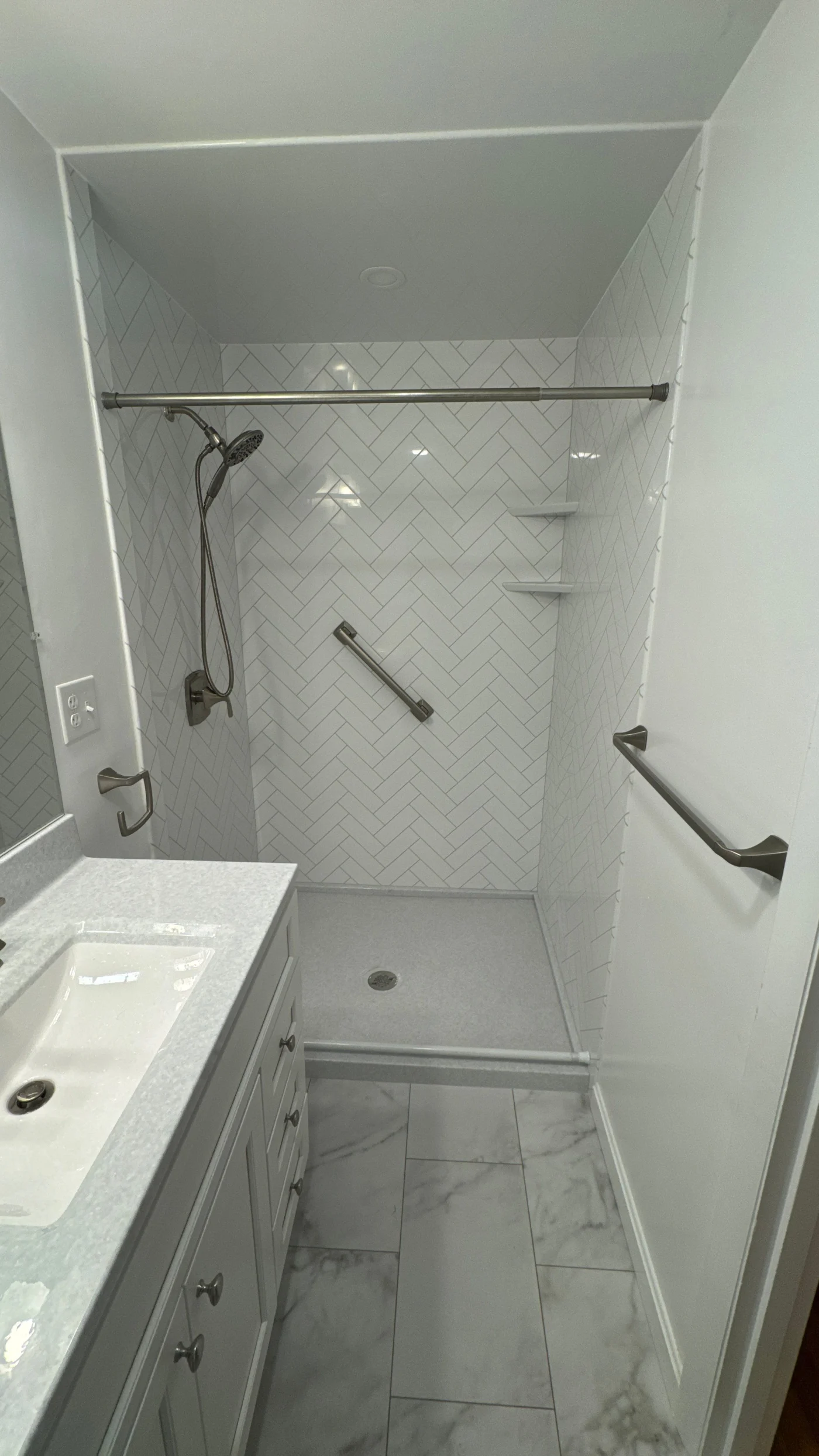 This pristine bathroom remodel features a white single-sink vanity with a speckled countertop, situated across from a low-threshold walk-in shower with white herringbone tiling and a convenient safety grab bar.