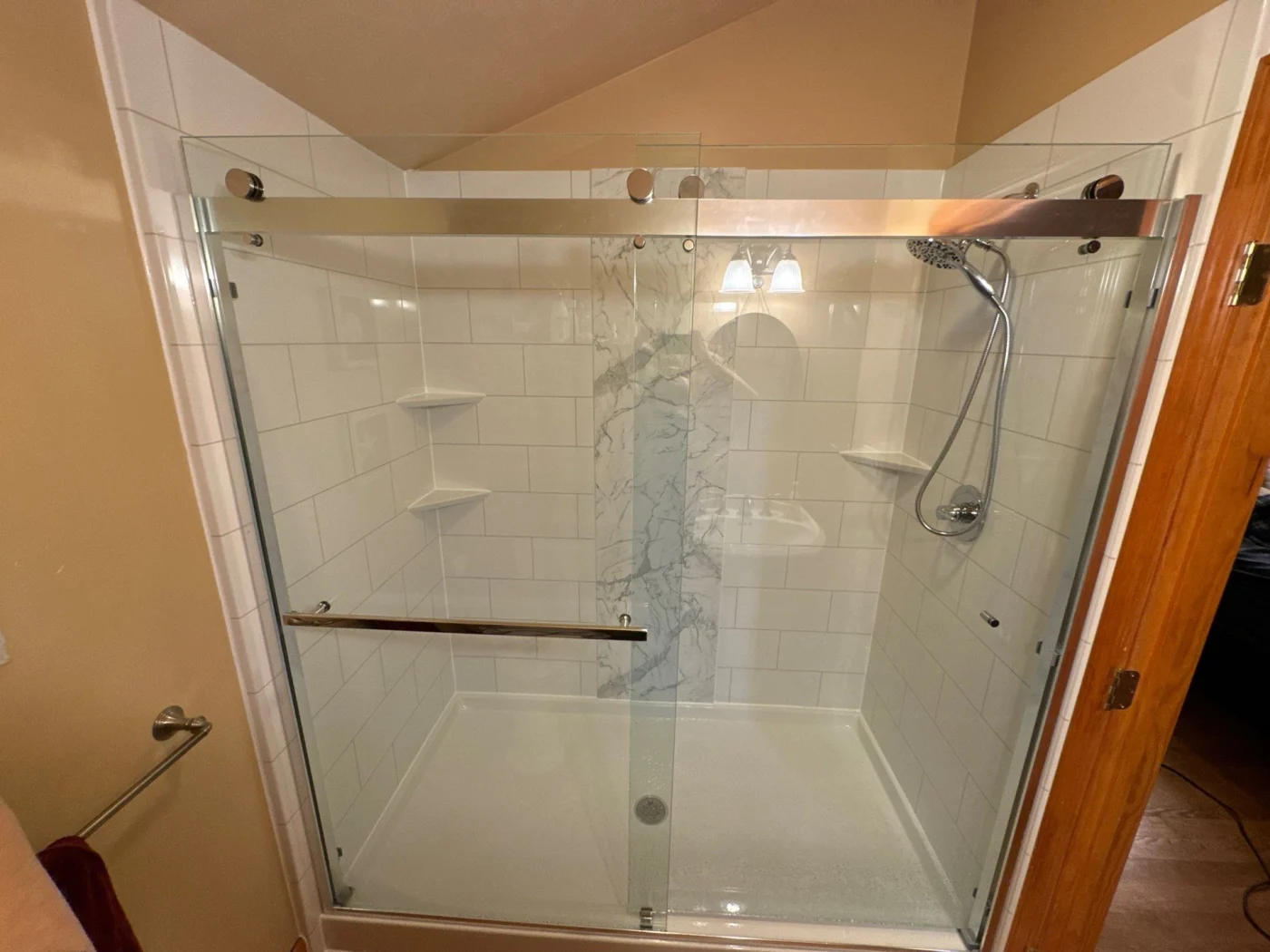 A modern walk-in shower features white subway tile walls accented by a central vertical marble strip, sliding glass doors, chrome hardware, and integrated corner shelves.