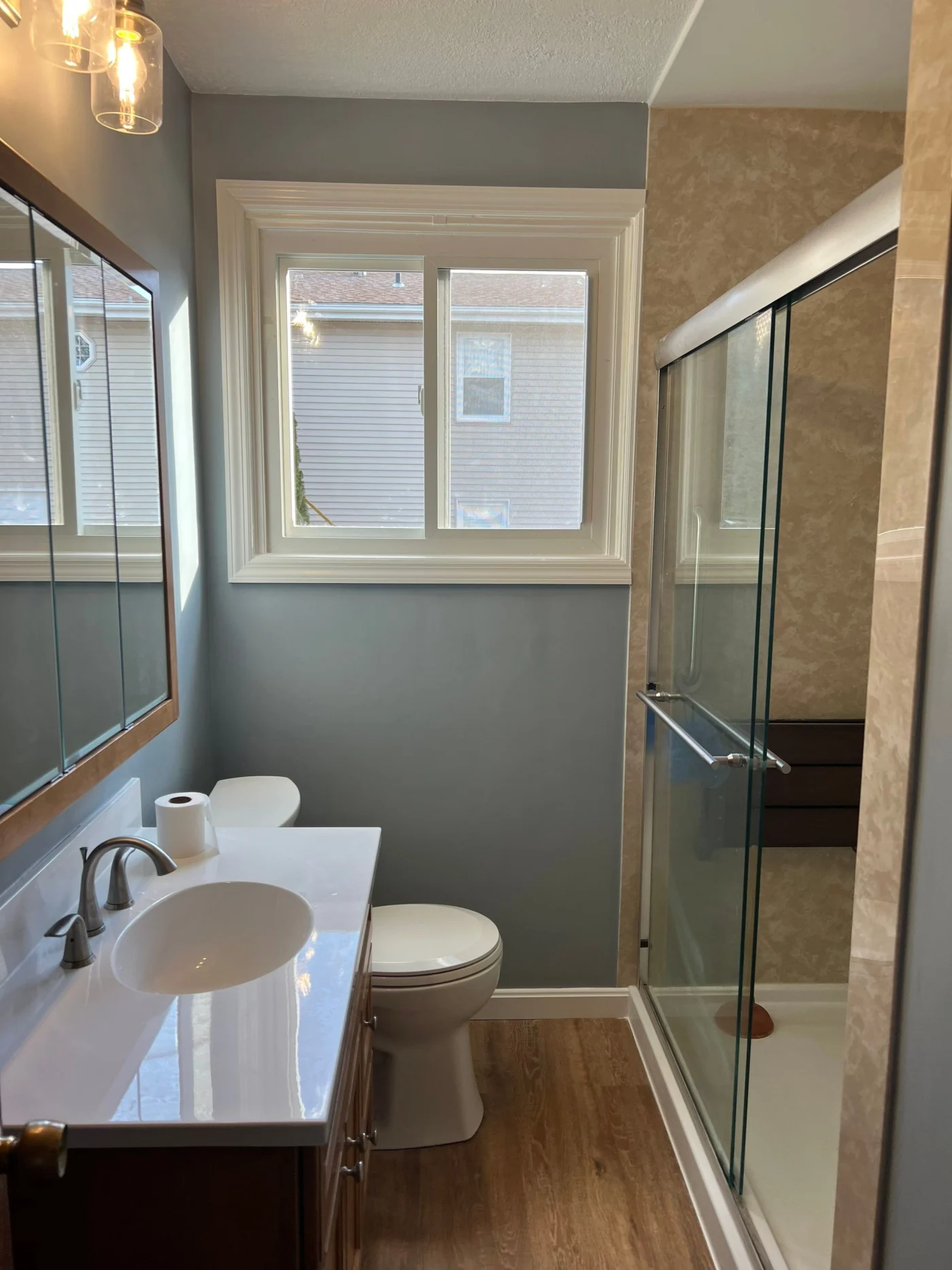 A remodeled bathroom features blue-gray walls, a wood-framed vanity with a white integrated sink, a large central window, and a glass-enclosed walk-in shower with beige textured walls and a wooden fold-down bench.