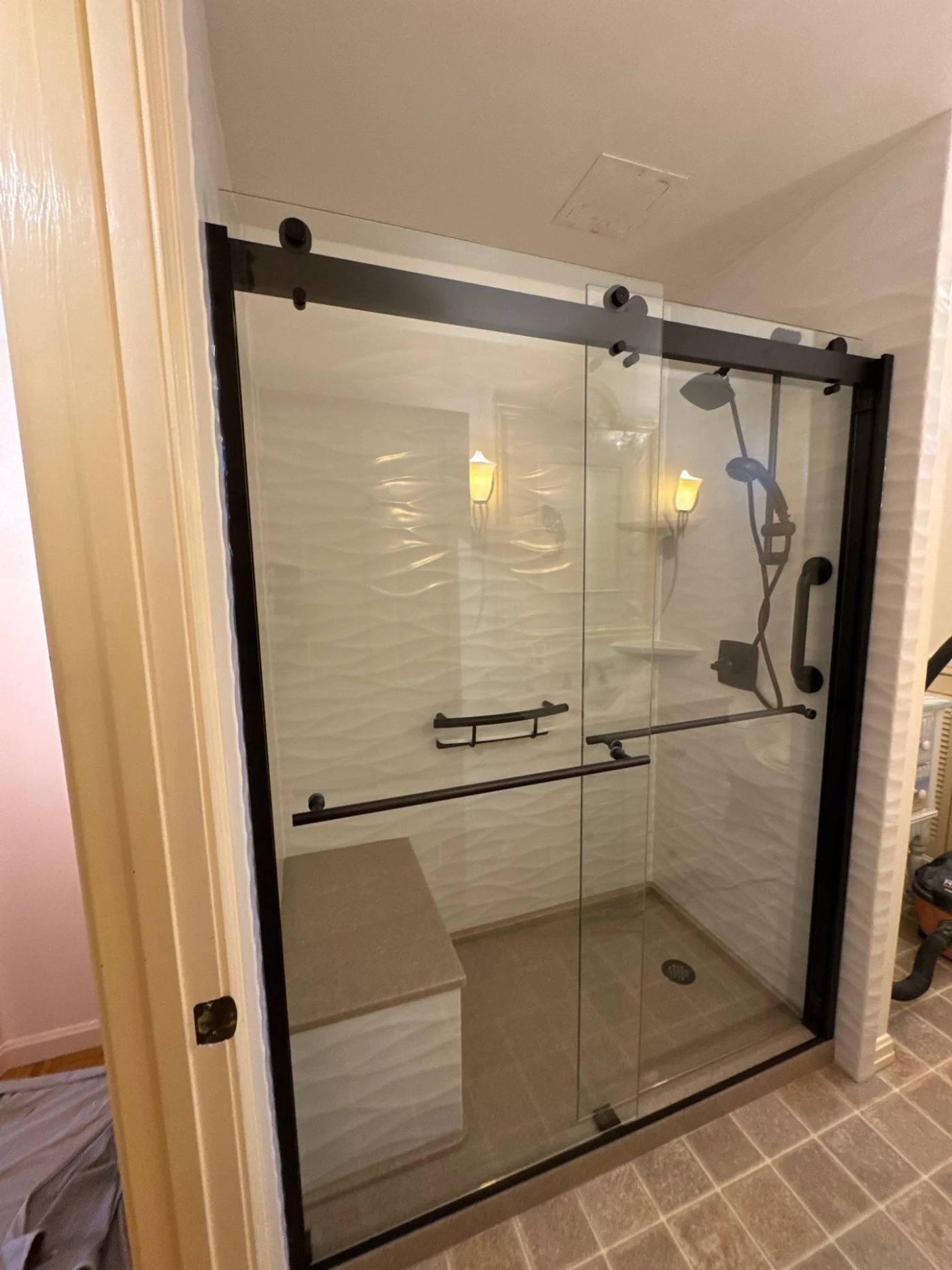 A walk-in shower with a brown base, white walls, a brown seat, black fixtures, and a glass door with a black frame.