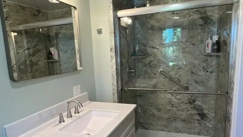 Remodeled bathroom with a marble-look walk-in shower, sliding glass doors, grab bars, and a white vanity.