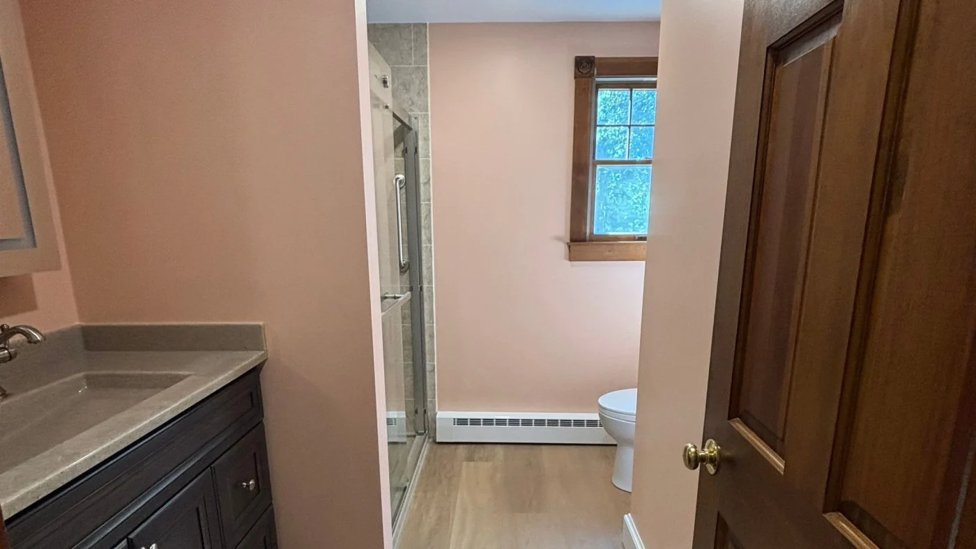 Remodeled bathroom with a dark vanity, walk-in shower, wood-look flooring, and natural wood window trim.