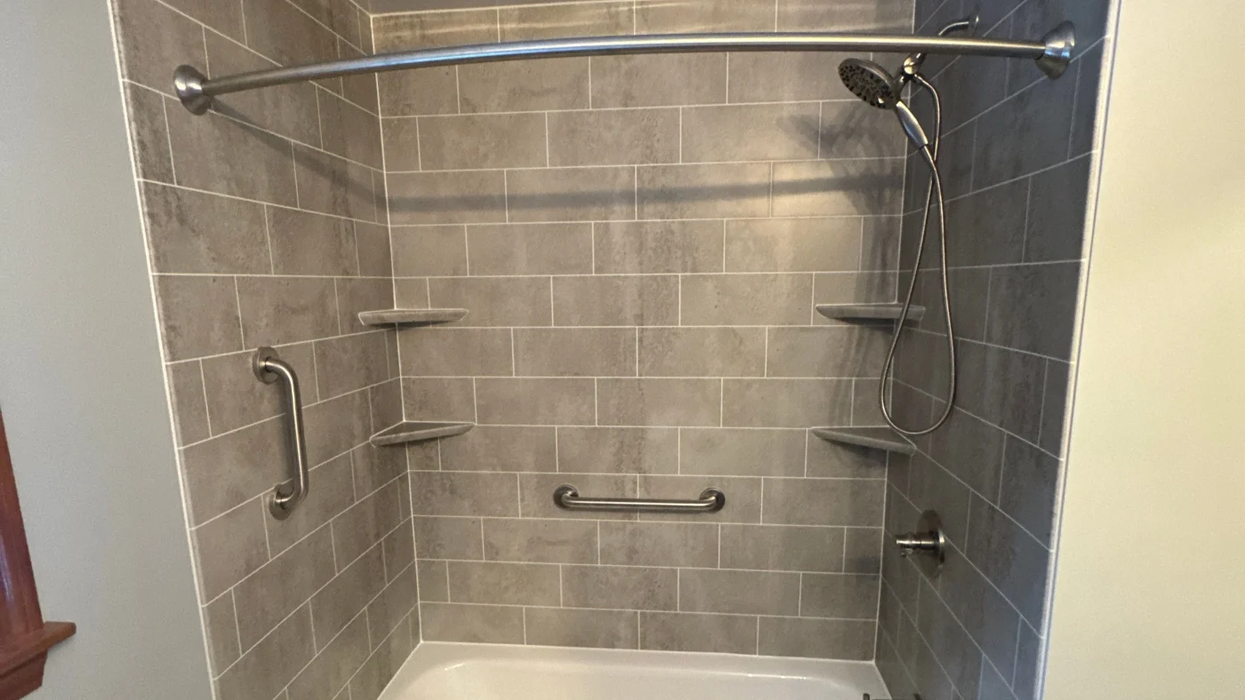 Gray tiled walk-in shower with grab bars, corner shelves, and a handheld showerhead.