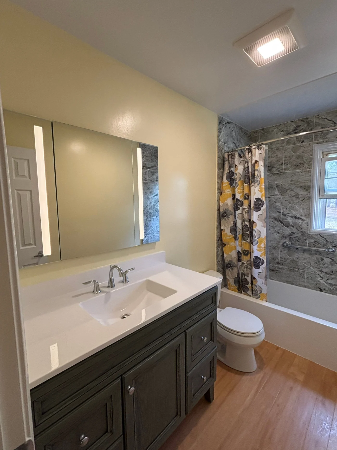 A bright bathroom features light yellow walls, a charcoal gray vanity with a white integrated sink, a large backlit mirror, and an alcove bathtub with gray marbled tiling and a yellow and gray floral shower curtain.