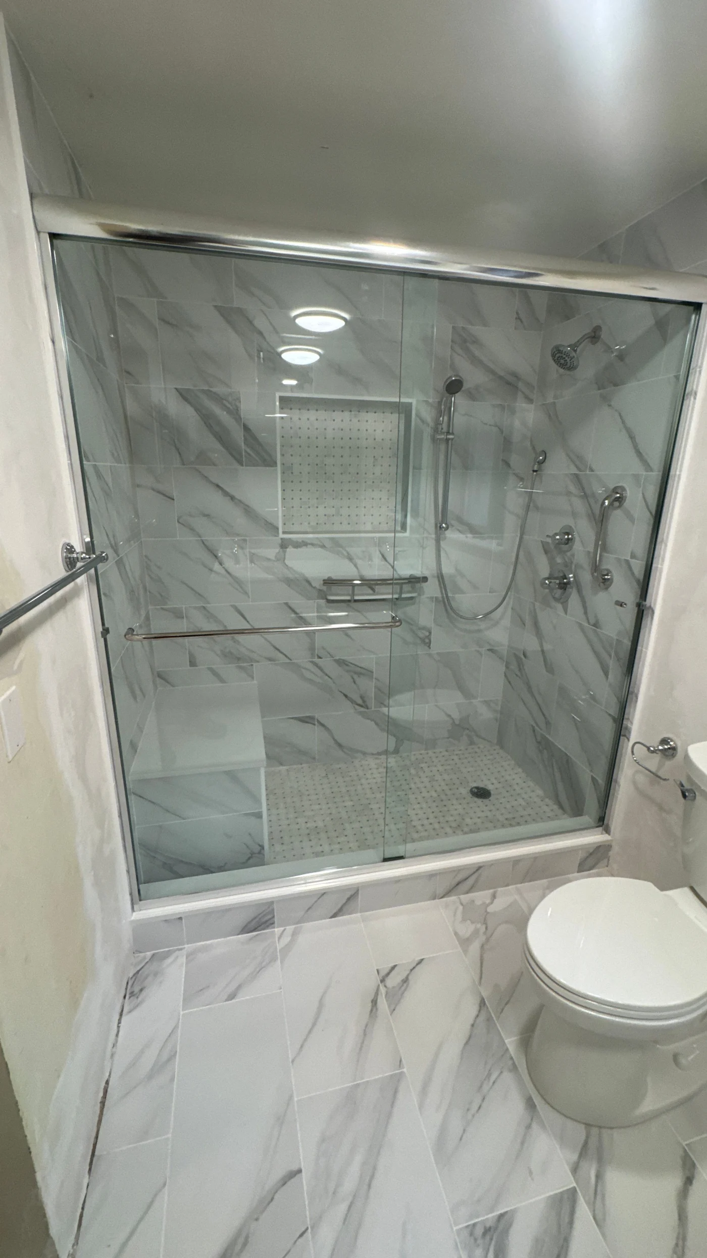 A bathroom with a white and gray tile floor, a white toilet, and a walk-in shower with silver fixtures and a sliding glass door.