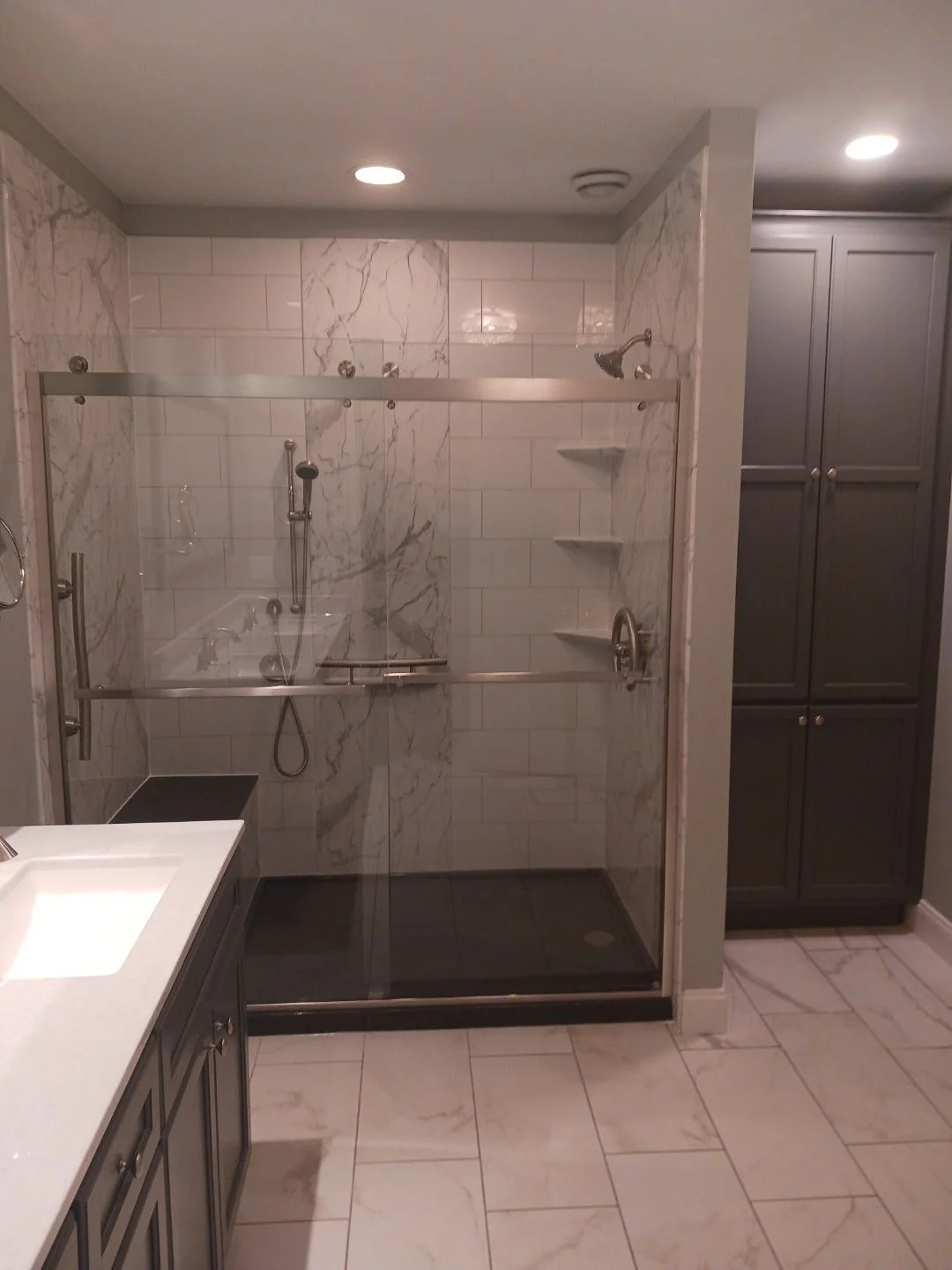 A white bathroom with a tile floor, a wooden vanity with a white marble countertop, a walk-in shower with white tile walls, a black base, and a black bench, and gray cabinets.