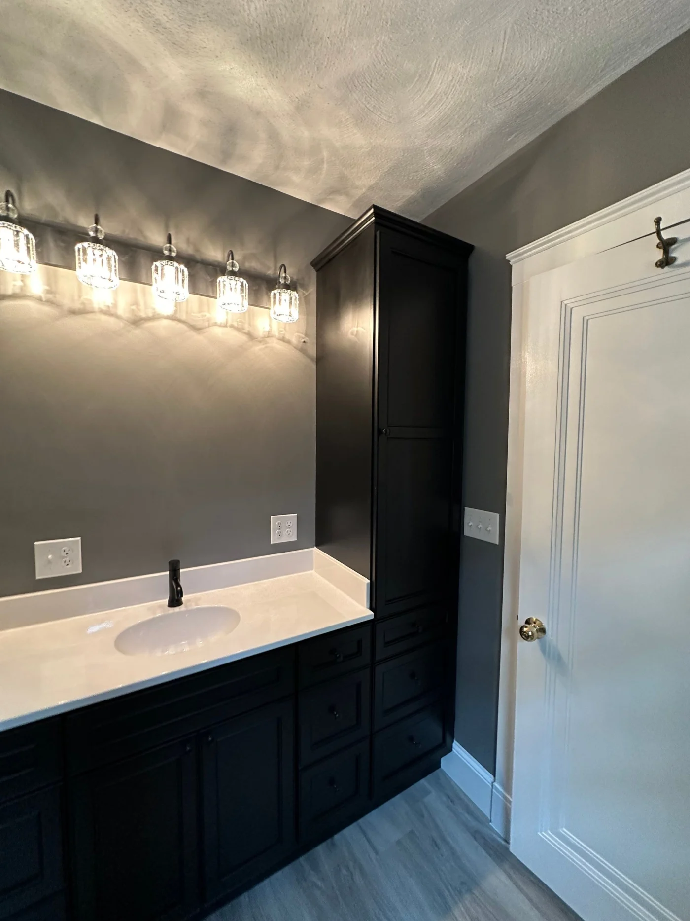 A gray bathroom with a white door, gray flooring, a black vanity with a white marble sink, a black faucet, and overhead lighting.