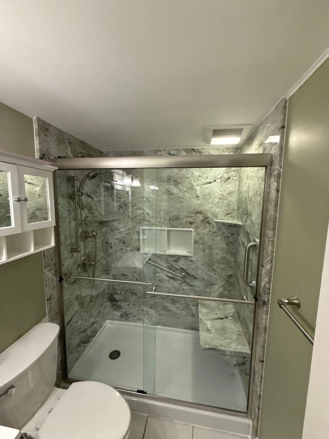 A gray walk-in shower with a white base, a gray bench, silver fixtures, shelves, and a sliding glass door.