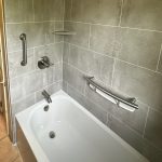 A white bathtub with a faucet, handle, and grab bar with a gray wall and a shelf.