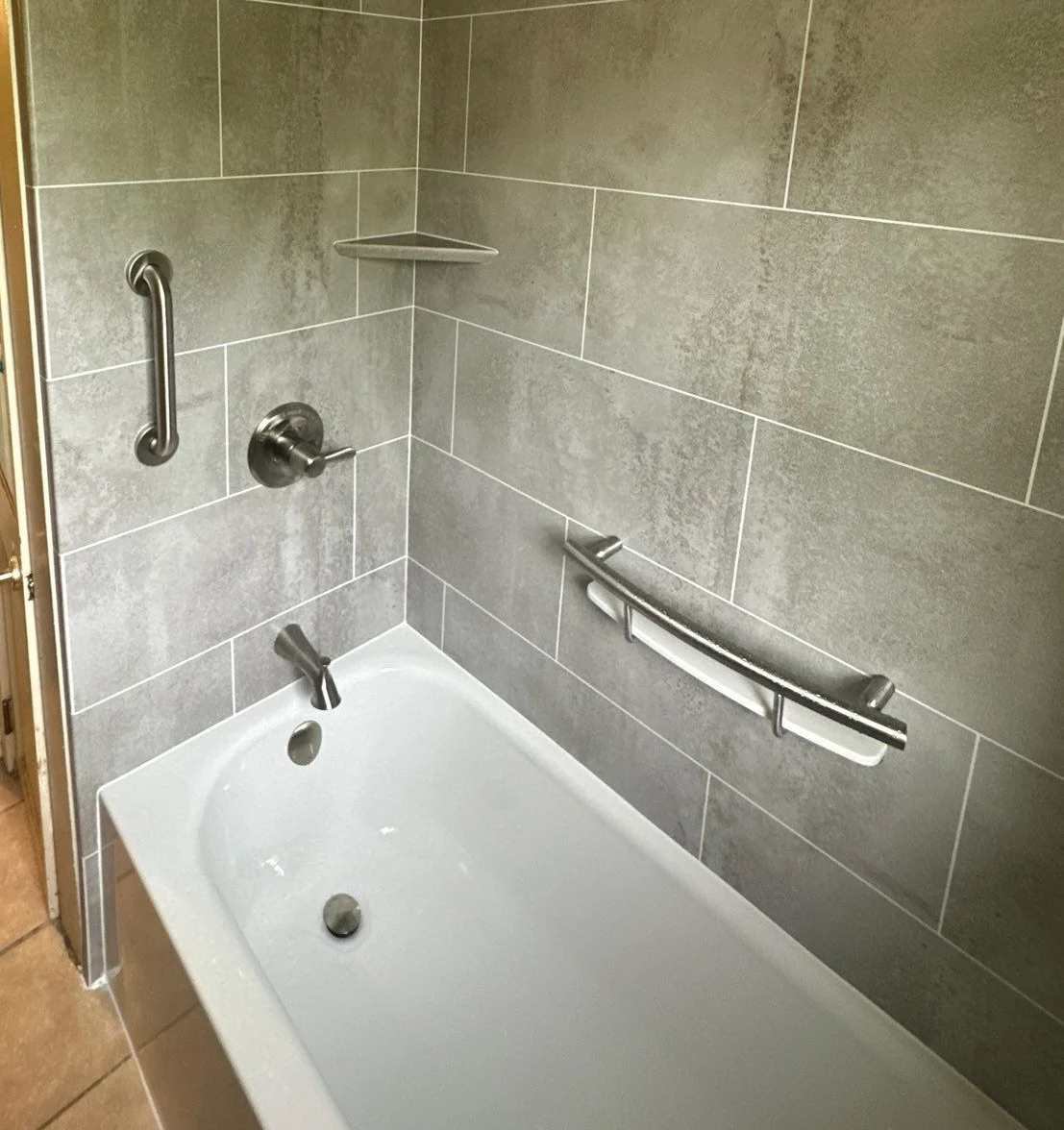 A white bathtub with a faucet, handle, and grab bar with a gray wall and a shelf.